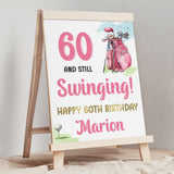 A personalized woman Golfer 60th Birthday sign with a golf bag and golf ball on a tee. 60 and Still Swinging! Happy 60th Birthday editable sign template. Pink and Gold.